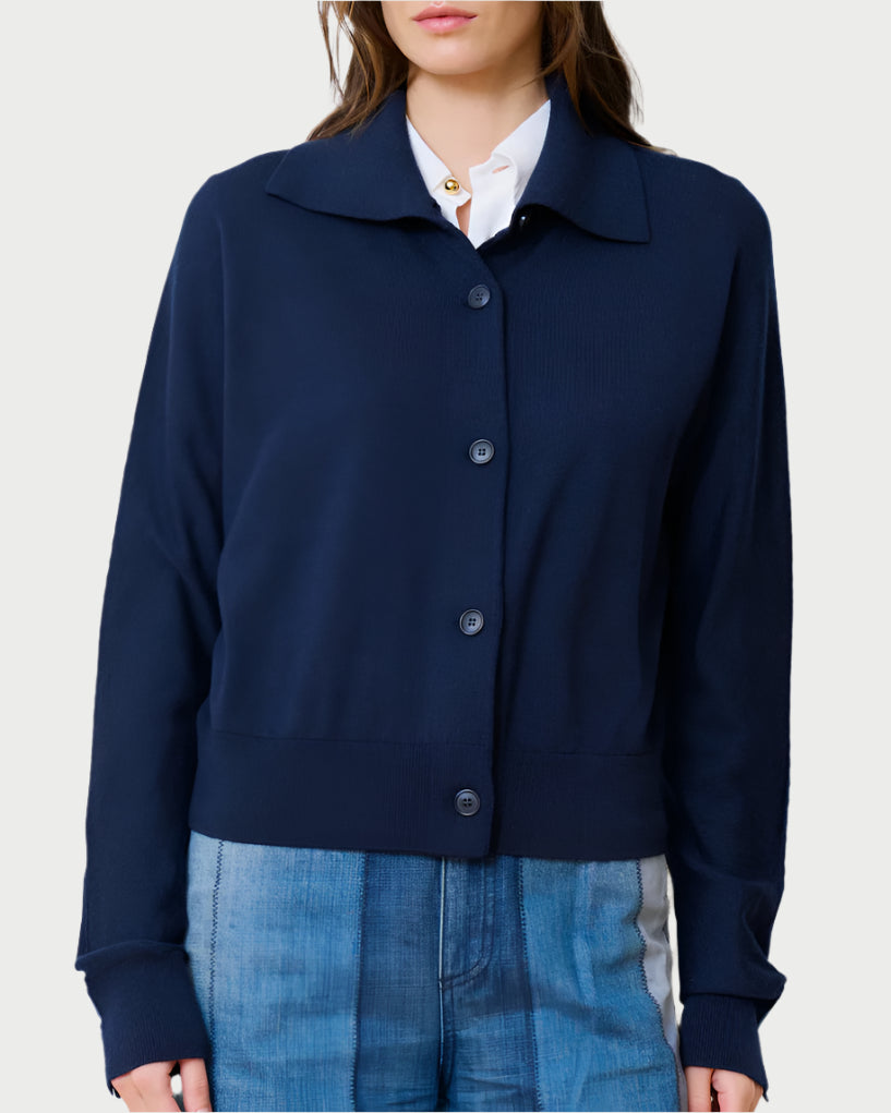 A woman wears the WE-AR4 Social Cardigan Navy, a Merino wool button-up, over a gold-detailed white collared shirt. She complements it with relaxed-fit denim pants in shades of blue, against a plain backdrop.