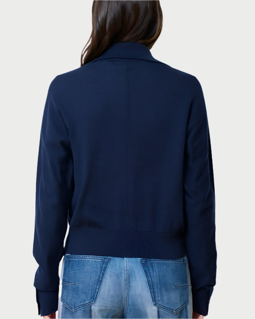 A person with long brown hair faces away, wearing a navy Social Cardigan by WE-AR4 made of Merino wool, paired with blue jeans. The background is a plain, light-colored wall.