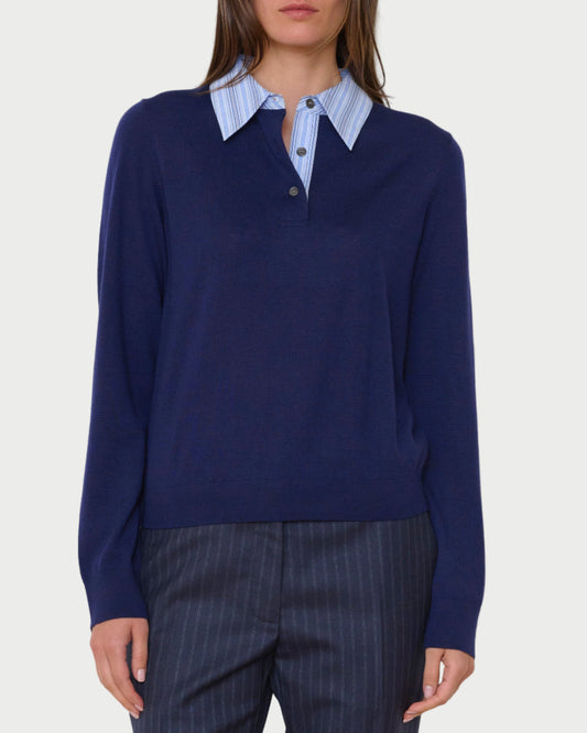 Person wearing a navy sweater over a striped shirt with dark pants against a plain background