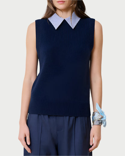 Against a plain backdrop, an individual dons the School Vest Navy by WE-AR4, a sleeveless sweater made from upcycled merino wool. A white-and-blue striped collared shirt complements their long brown hair and light blue scarf on the wrist—an epitome of sustainable fashion.