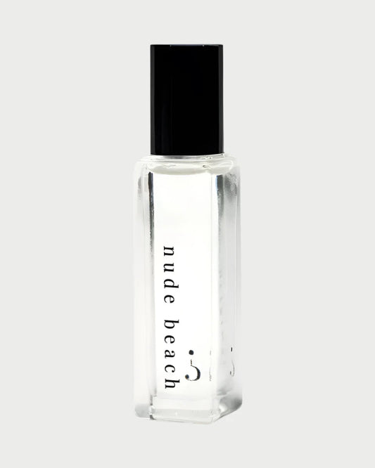 A Riddle Oil - Nude Beach 8ml bottle, made of clear glass and sealed with a black cap, contains a transparent liquid. The label features the words "nude beach" in lowercase letters. The design is minimalist and sleek, showcasing the trending Nude Beach scent by Riddle Oil.