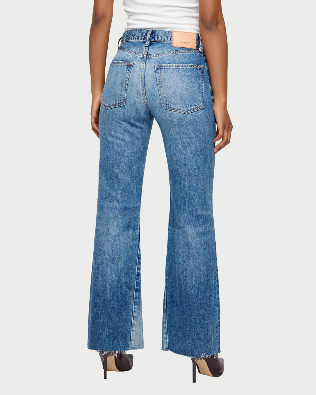 A person is seen from the back wearing the Moussy Asheville Flare Jean by Moussy, a pair of vintage-inspired low-rise flare jeans with frayed hems, crafted in Japan. The jeans feature large back pockets and a tan label on the waistband. The outfit is completed with dark heels and a white top.