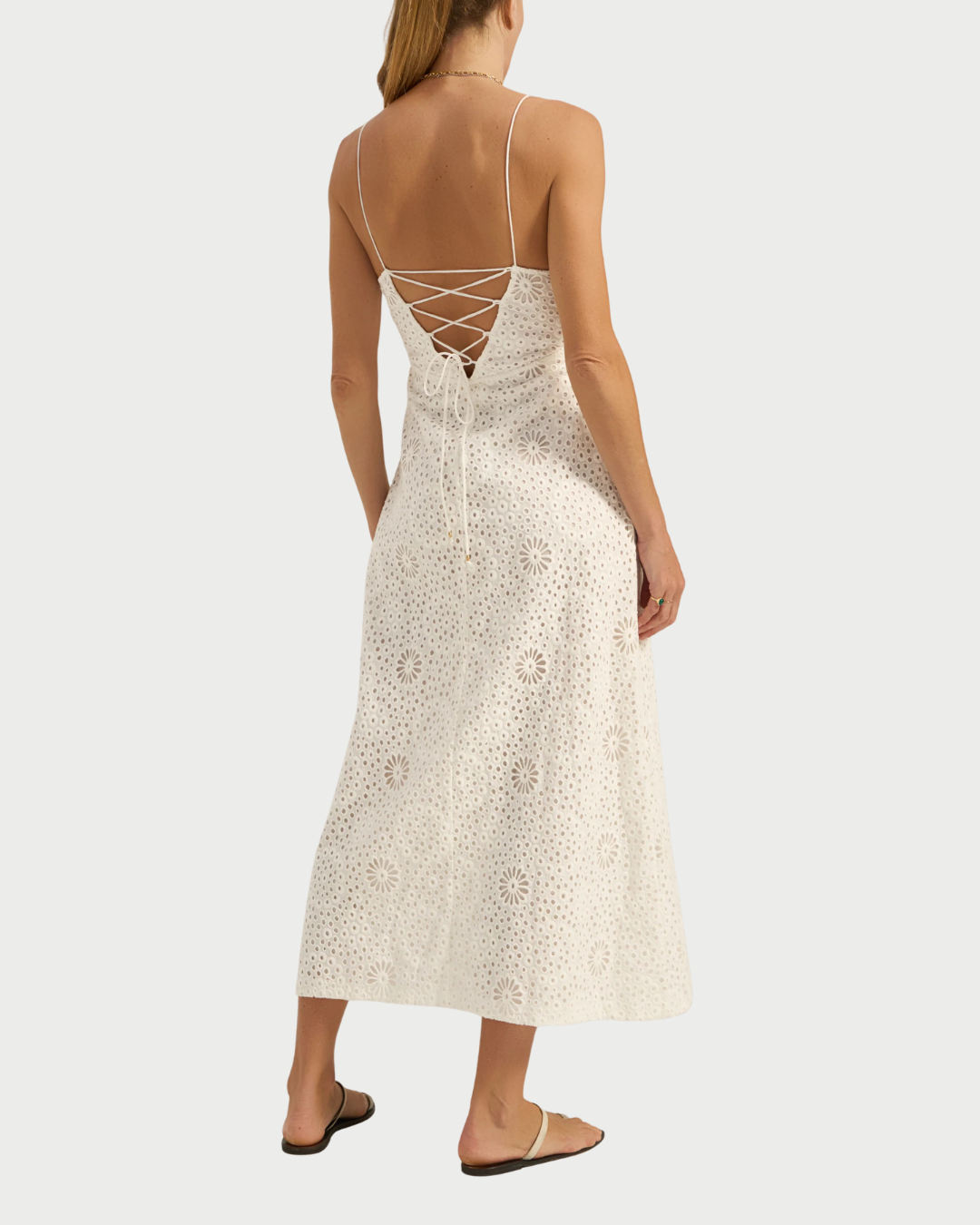 A woman wearing the Ilio Nema Ino Dress, a white eyelet midi dress with lace-up back and spaghetti straps by Ilio Nema, pairs it with flat sandals and a necklace for an effortlessly breezy summer look.