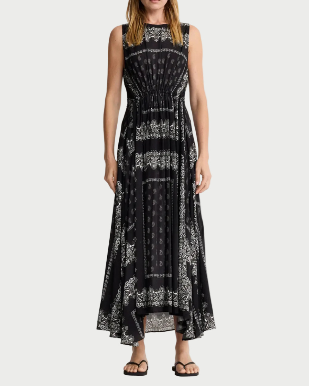 A woman wears the Smythe Drawstring Maxi Dress by Smythe, featuring a black bandana print with white paisley details, an asymmetrical hem, and eco-conscious viscose fabric, styled with black sandals.