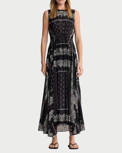 A woman wears the Smythe Drawstring Maxi Dress by Smythe, featuring a black bandana print with white paisley details, an asymmetrical hem, and eco-conscious viscose fabric, styled with black sandals.