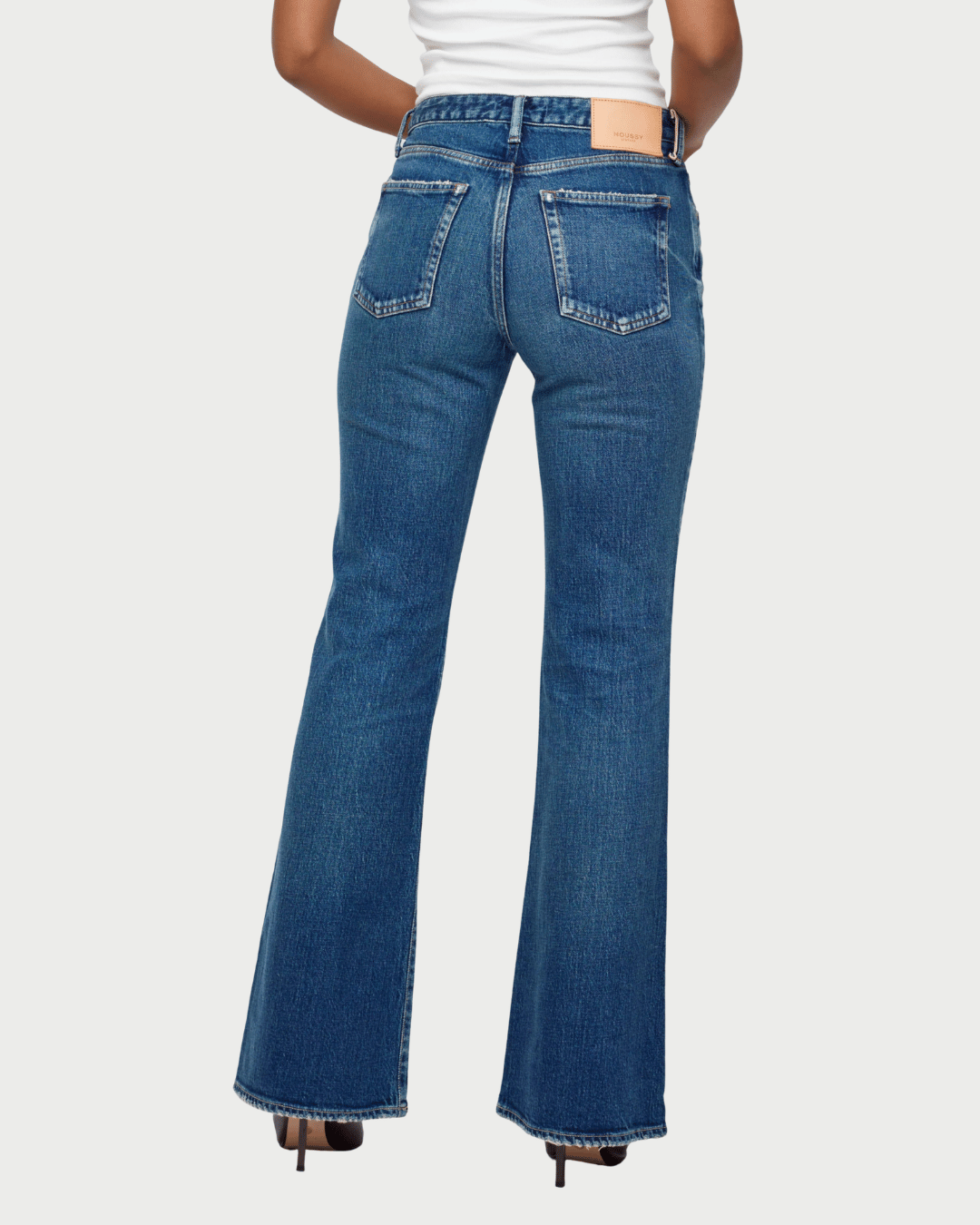 Rear view of a model wearing blue denim flare jeans with a white top and black heels, set against a light gray studio background.