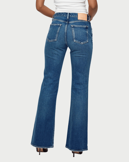 Rear view of a model wearing blue denim flare jeans with a white top and black heels, set against a light gray studio background.