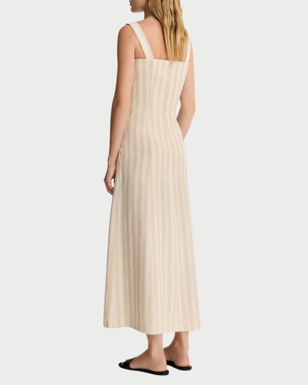 A woman wearing the Smythe Picnic Dress by Smythe—a sleeveless, ankle-length cream and beige striped dress with adjustable straps—stands facing away, her straight blonde hair and black flat sandals against a plain white background.