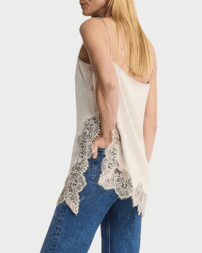 A woman wearing a cream-colored spaghetti-strap camisole top with intricate lace trim along the hem, paired with blue jeans, shown from a side-back angle against a light gray background.