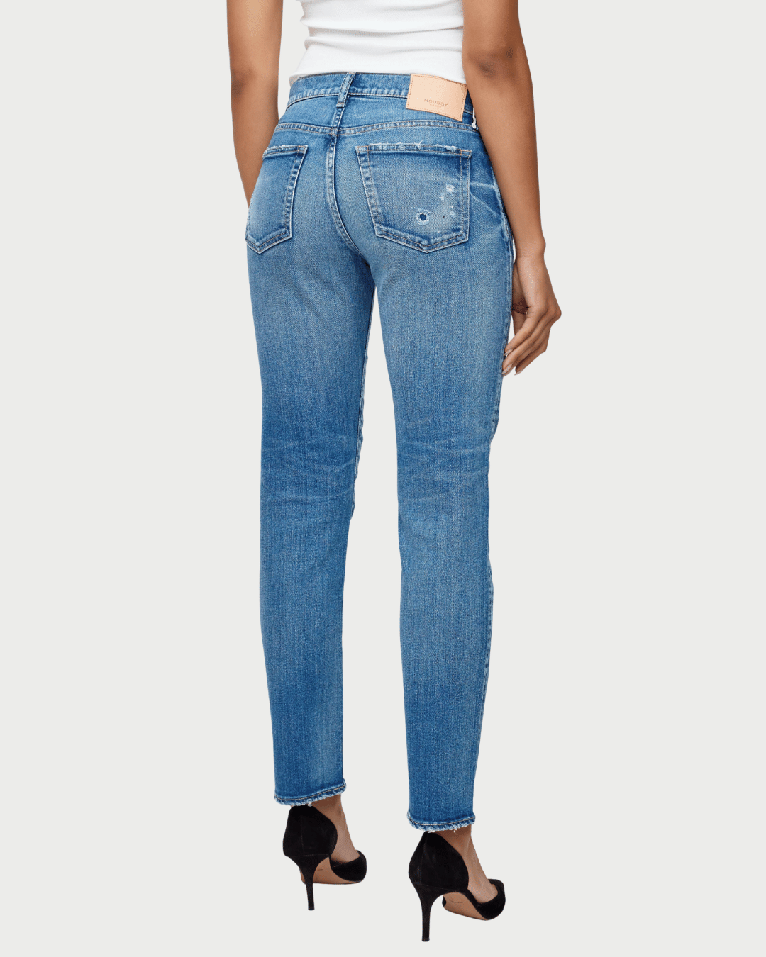 Rear view of a model wearing medium-wash blue skinny jeans with a white top and black high heels, shown against a light gray studio background.