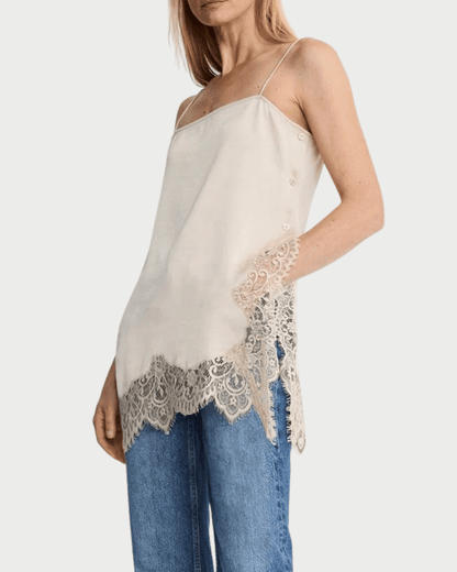 A woman wearing the Smythe Side Button Cami by Smythe—a white cami with lace trim and thin straps—paired with blue jeans, stands against a plain background, showcasing this effortless layering essential for any wardrobe.