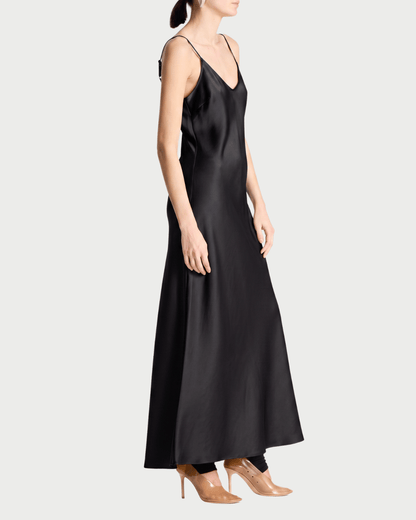 A model wearing a black satin slip dress with thin spaghetti straps and a V-neckline, paired with clear high-heeled pumps, shown from a side profile against a light gray background.