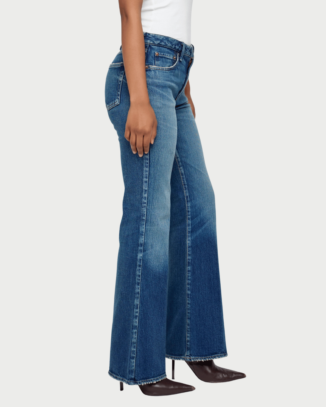 A side-profile view of a model wearing medium-wash blue flare jeans with frayed hems, paired with a white top and dark brown pointed-toe heels, against a light gray studio background.
