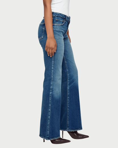 A side-profile view of a model wearing medium-wash blue flare jeans with frayed hems, paired with a white top and dark brown pointed-toe heels, against a light gray studio background.