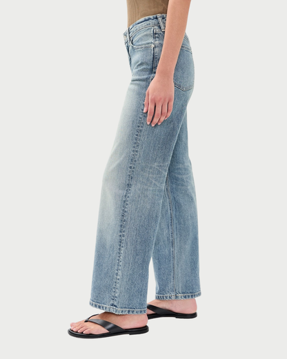 A side profile view of a person wearing light-wash blue wide-leg jeans and black flat sandals against a light gray studio background.