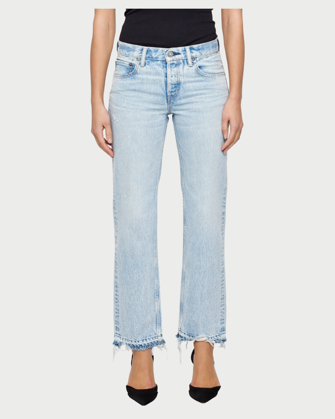 A woman wears the Moussy Barrington Straight Low Rise Jean by Moussy, light blue straight-leg jeans made from Japanese denim with frayed hems, paired with a black tank top and black pointed-toe heels.
