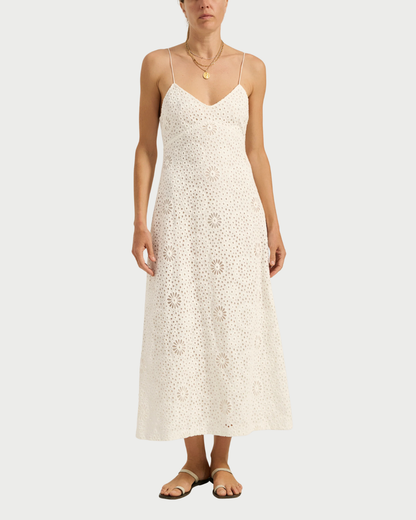 A woman models the Ilio Nema Ino Dress—a sleeveless, white eyelet maxi with thin straps—paired with sandals and layered gold necklaces by Ilio Nema, creating a chic summer look against a plain background.
