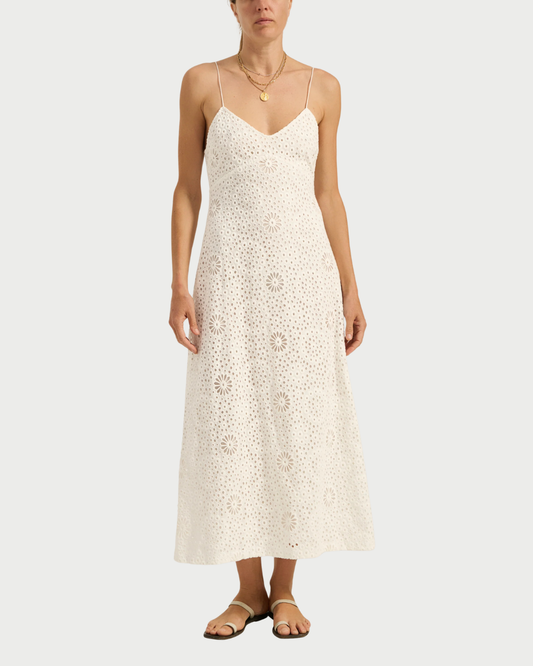 A woman models the Ilio Nema Ino Dress—a sleeveless, white eyelet maxi with thin straps—paired with sandals and layered gold necklaces by Ilio Nema, creating a chic summer look against a plain background.