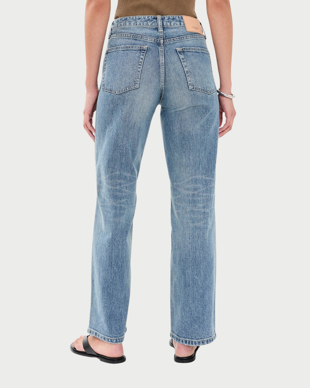 Rear view of a person wearing light-wash blue denim jeans with a straight-leg fit, paired with black sandals and a brown top, against a light gray studio background.