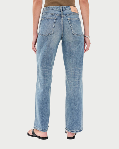 Rear view of a person wearing light-wash blue denim jeans with a straight-leg fit, paired with black sandals and a brown top, against a light gray studio background.