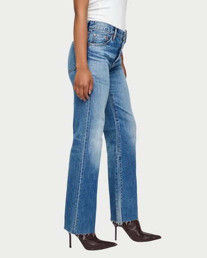 Wearing the Moussy Asheville Flare Jean by Moussy in vintage-inspired denim blue with a raw hem, styled with a white top and pointed dark brown high-heeled boots, the person stands against a plain light background.