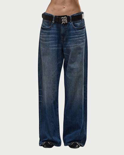 Wearing R13 D'Arcy Loose Jean by R13 in a vintage-inspired dark blue wash, paired with a black belt featuring a large decorative “B” buckle; midriff exposed and shoes visible beneath the wide-leg hem.