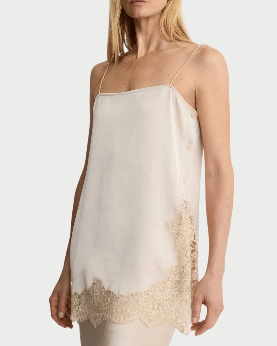 A woman wears the Smythe Side Button Cami in a cream color, featuring thin straps, delicate lace trim on the hem and side, and elegant button details along the side. Designed by Smythe, this cami offers a loose, graceful fit—making it a versatile layering essential.