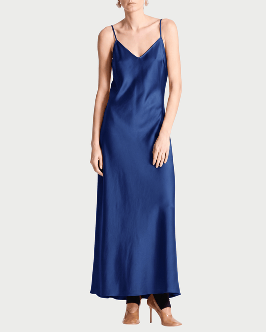 A woman modeling a long, royal blue satin slip dress with thin spaghetti straps and a V-neckline, styled with nude high heels against a light gray studio background.