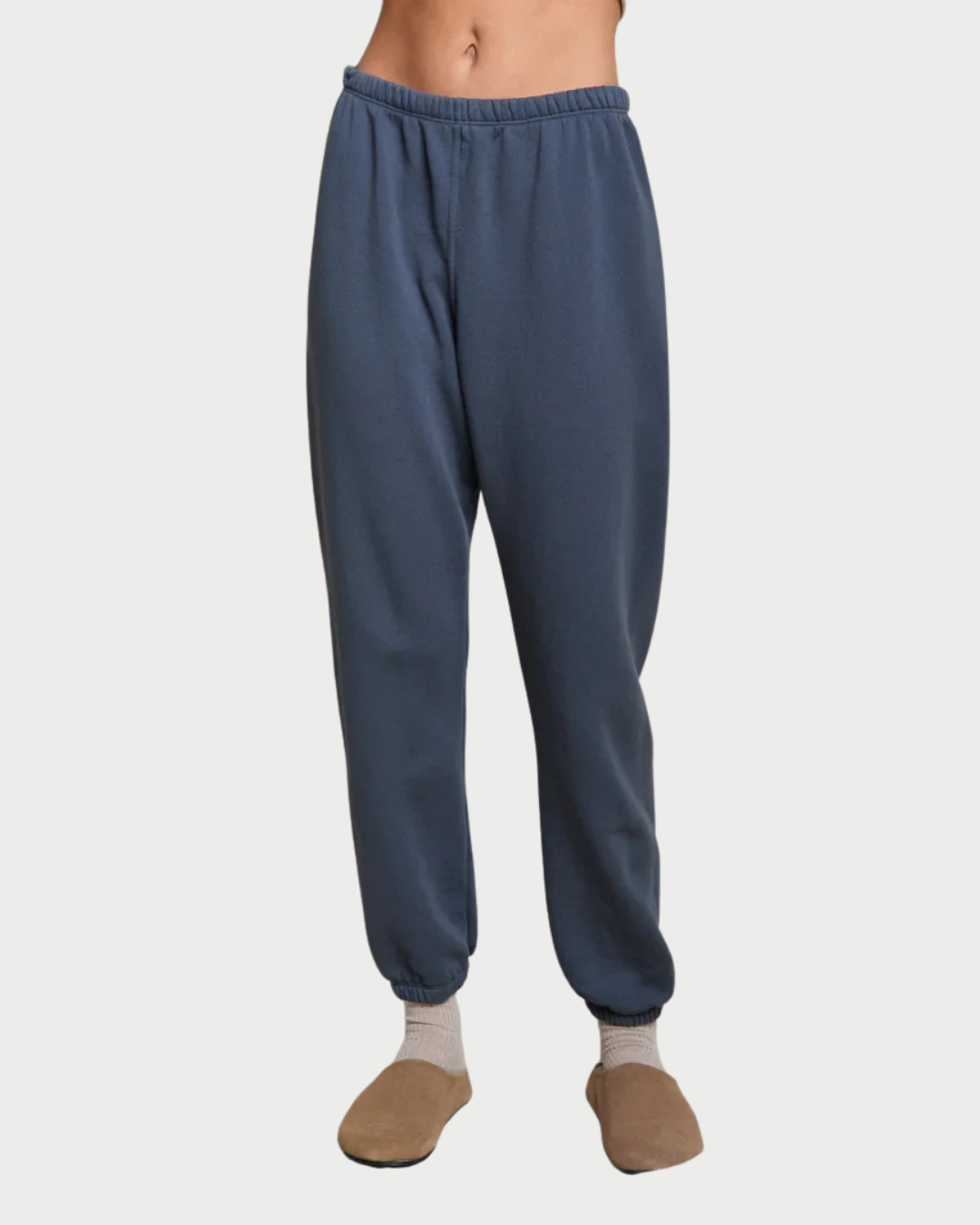A person wearing slate blue jogger sweatpants with an elastic waistband and cuffs, paired with beige socks and tan slippers, shown against a light gray studio background.