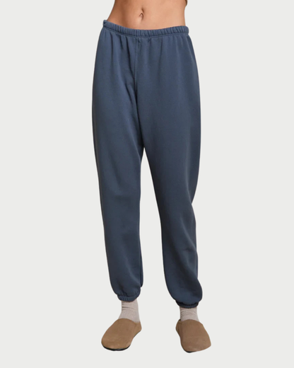 A person wearing slate blue jogger sweatpants with an elastic waistband and cuffs, paired with beige socks and tan slippers, shown against a light gray studio background.