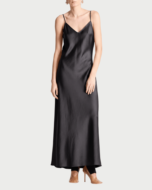 A woman wearing the Saint Art Olivia Slip Dress by Saint Art NY—a long, black satin slip dress with adjustable straps—and tan heeled shoes, standing against a plain white background.