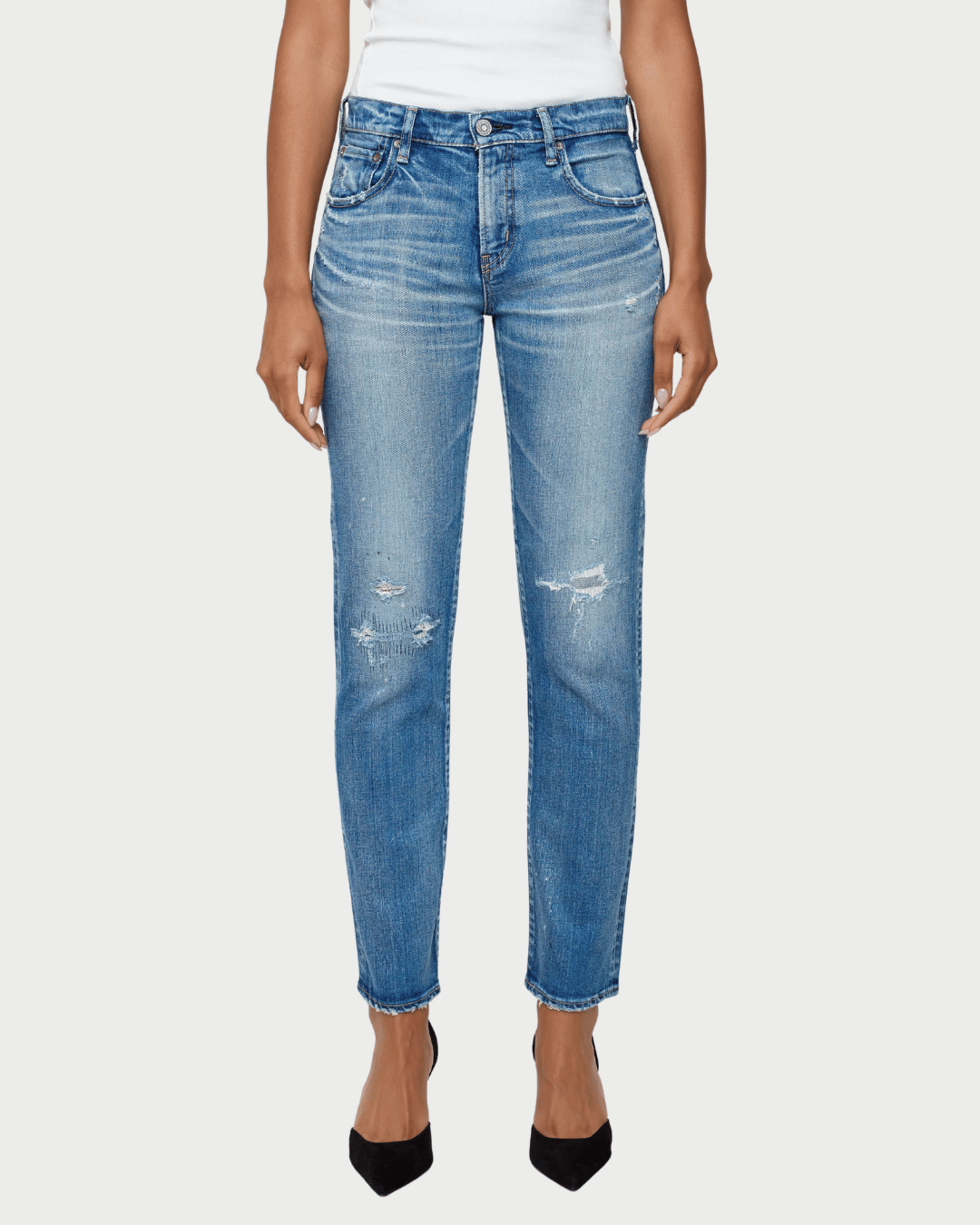 A model wearing a pair of medium-wash blue denim jeans with distressed details on the thighs and knees, paired with a white top and black heels, against a light gray background.