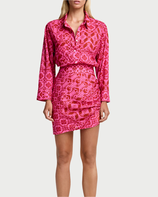 A person stands facing forward, wearing the Smythe Mini Shirt Dress in hot pink with a matching pattern. They have long, straight hair and brown slippers, set against a plain white background.
