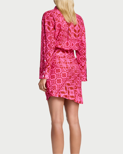 A person with long blonde hair, facing away, wears the Smythe Mini Shirt Dress in bright hot pink with long sleeves and beige slip-on shoes against a white background.