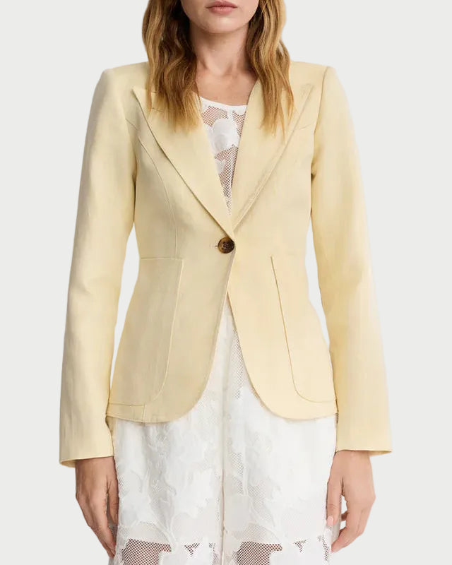 A woman stands against a plain background wearing the Smythe Patch Pocket Duchess Blazer in light yellow over a white lace dress. The tailored Smythe blazer features leather elbow patches, patch pockets, a single button, and notched lapels.