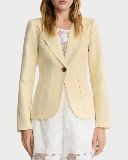 A woman stands against a plain background wearing the Smythe Patch Pocket Duchess Blazer in light yellow over a white lace dress. The tailored Smythe blazer features leather elbow patches, patch pockets, a single button, and notched lapels.