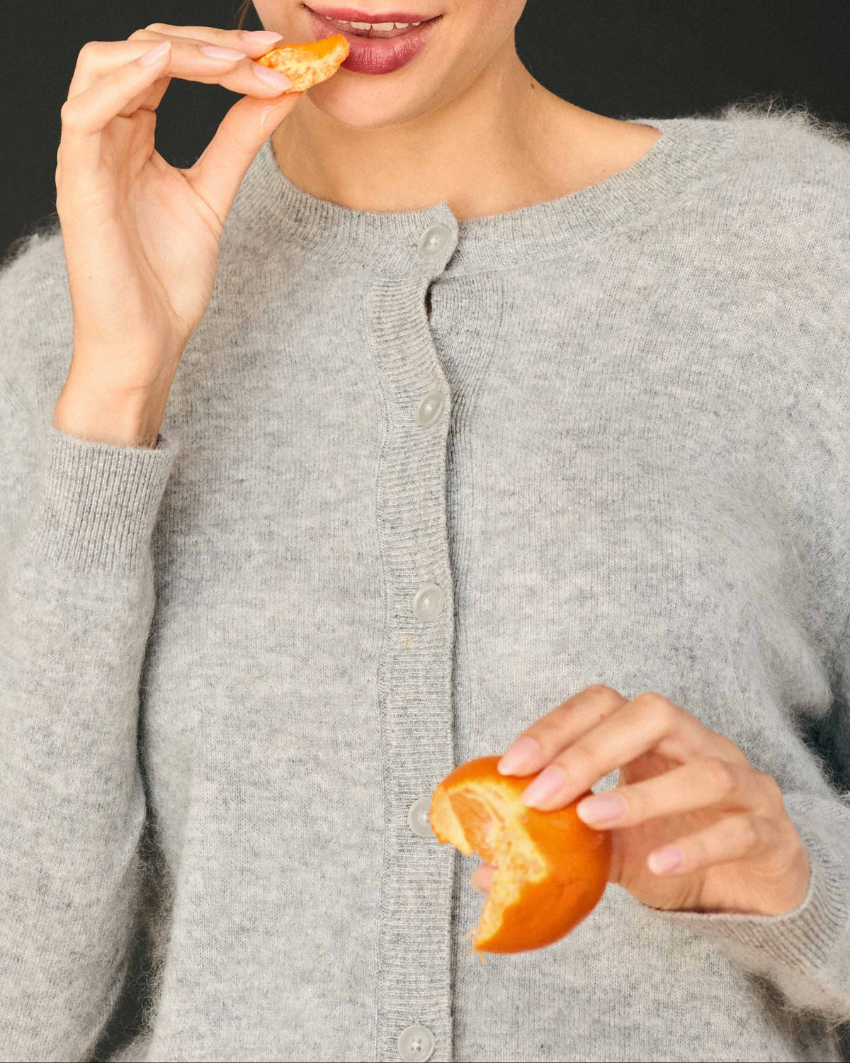 A person wearing an oversized, grey Jumper 1234 Brushed Cardigan eats a segment of a mandarin orange.