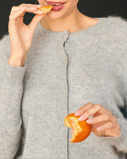 A person wearing an oversized, grey Jumper 1234 Brushed Cardigan eats a segment of a mandarin orange.