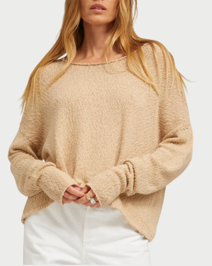 A woman with long, blonde hair wears the SPRWMN Off Shoulder Sweater by SPRWMN with white pants, standing against a plain light background and holding the hem of the sweater with both hands.