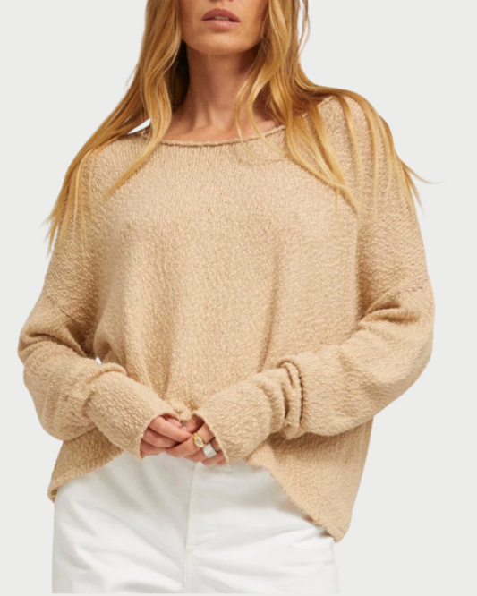 A woman with long, blonde hair wears the SPRWMN Off Shoulder Sweater by SPRWMN with white pants, standing against a plain light background and holding the hem of the sweater with both hands.