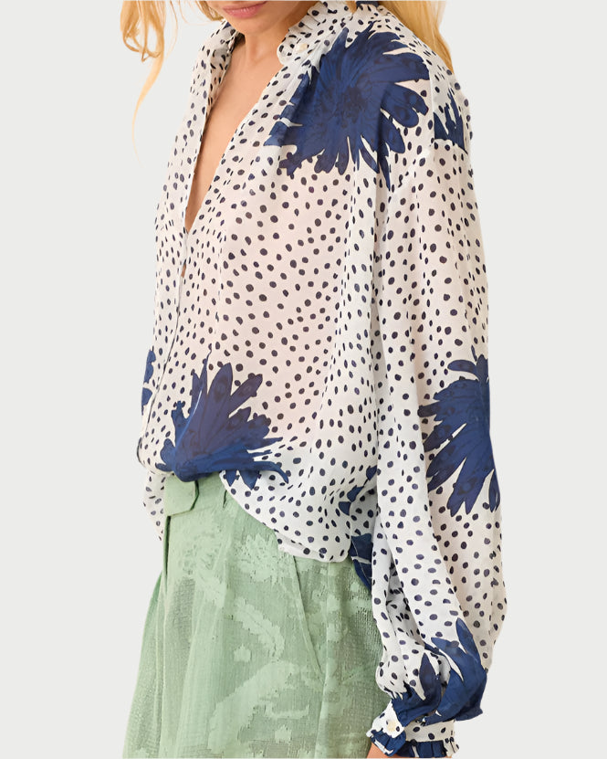 A woman wears the Poet Dahlia Blouse from Alix of Bohemia, featuring vintage-inspired blue floral patterns and small black dots. She pairs it with green textured pants as her blonde hair cascades gently against a plain white background.