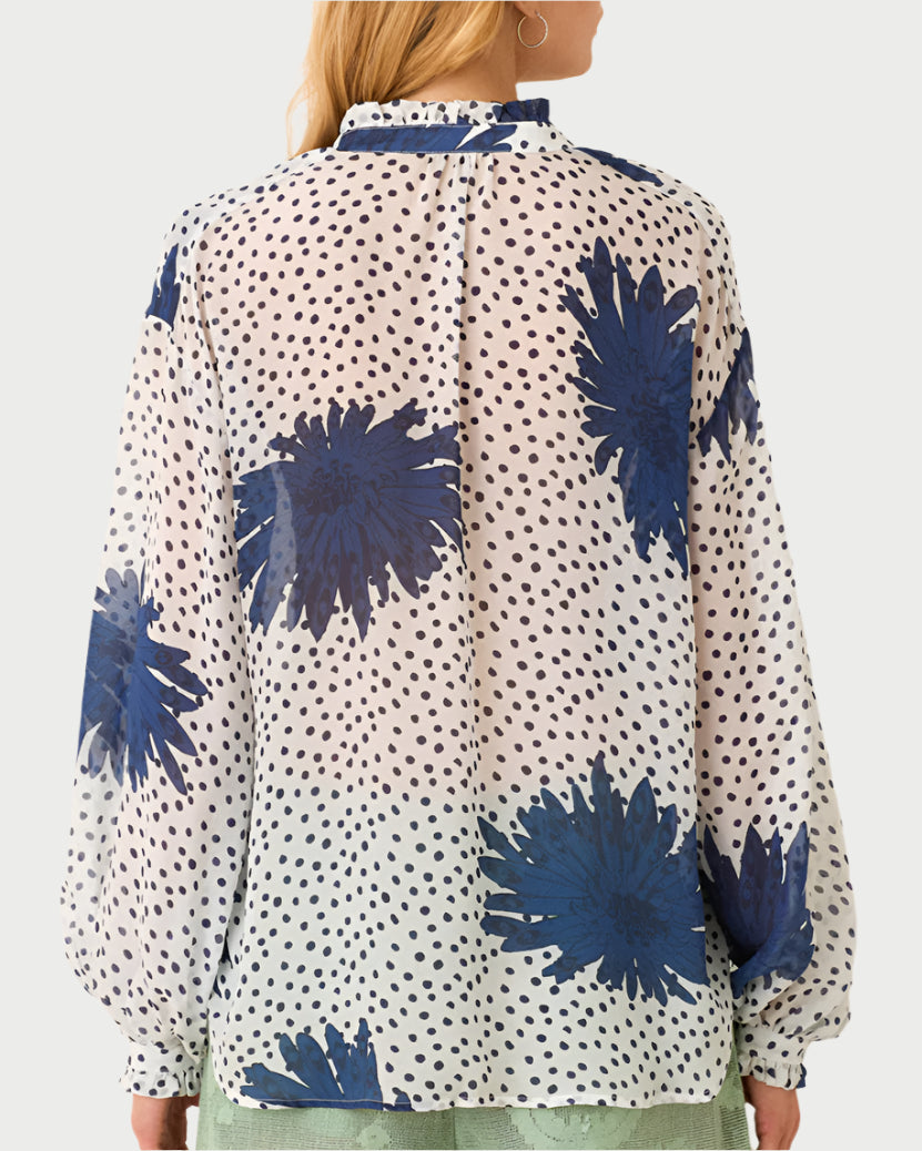 The Poet Dahlia Blouse by Alix of Bohemia showcases vintage blue floral patterns, a ruffled collar, and polka dots on high-collared long sleeves. Worn with a light green skirt, the look is completed as long hair cascades down an individual’s back while they face away from the camera.