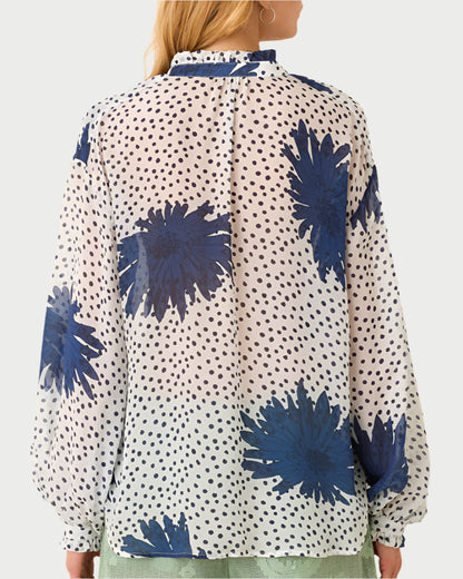 The Poet Dahlia Blouse by Alix of Bohemia showcases vintage blue floral patterns, a ruffled collar, and polka dots on high-collared long sleeves. Worn with a light green skirt, the look is completed as long hair cascades down an individual’s back while they face away from the camera.
