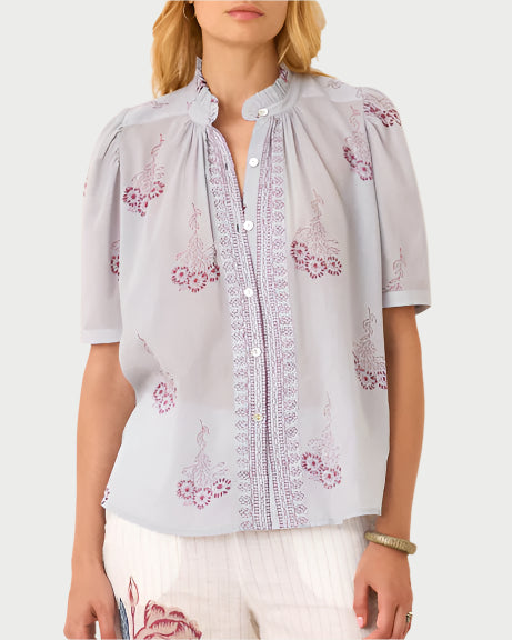 The woman wears the Pansy Sky Short Sleeve Shirt, an elegant cotton voile blouse by Alix of Bohemia, featuring intricate embroidery and maroon floral designs. It pairs seamlessly with white pants that have a subtle floral print near the waistline.