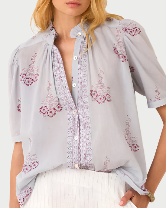 Wearing the Pansy Sky Short Sleeve Shirt by Alix of Bohemia, a woman showcases its light blue color with purple floral embroidery and puffed sleeves. Partially unbuttoned, she pairs it with white pants, her long blonde hair flowing freely and channeling bohemian charm.