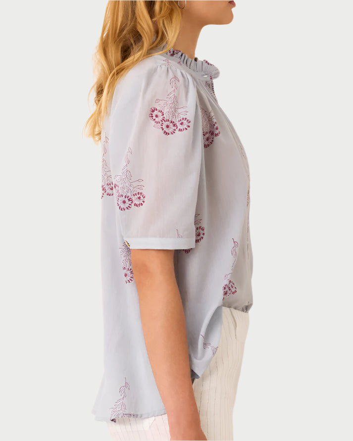 A person is seen from the side wearing Alix of Bohemia's Pansy Sky Short Sleeve Shirt, a light blue blouse with floral embroidery, and white pants. The cotton voile top has short sleeves and a ruffled collar that stand out beautifully against the off-white background.