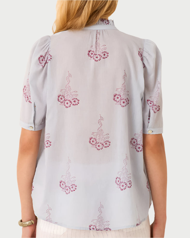 A woman with long hair and a bracelet stands with her back turned, wearing the Pansy Sky Short Sleeve Shirt from Alix of Bohemia. The vintage-inspired light blue cotton voile blouse features puffed sleeves, a red hand-block printed floral pattern, and subtle pleats at the back.