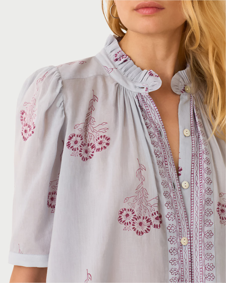 A woman wears the Pansy Sky Short Sleeve Shirt by Alix of Bohemia, featuring light gray cotton voile with maroon floral embroidery. The vintage-inspired design includes a ruffled collar, button-down front, and puffed sleeves; her upper body is visible with her face partially shown.