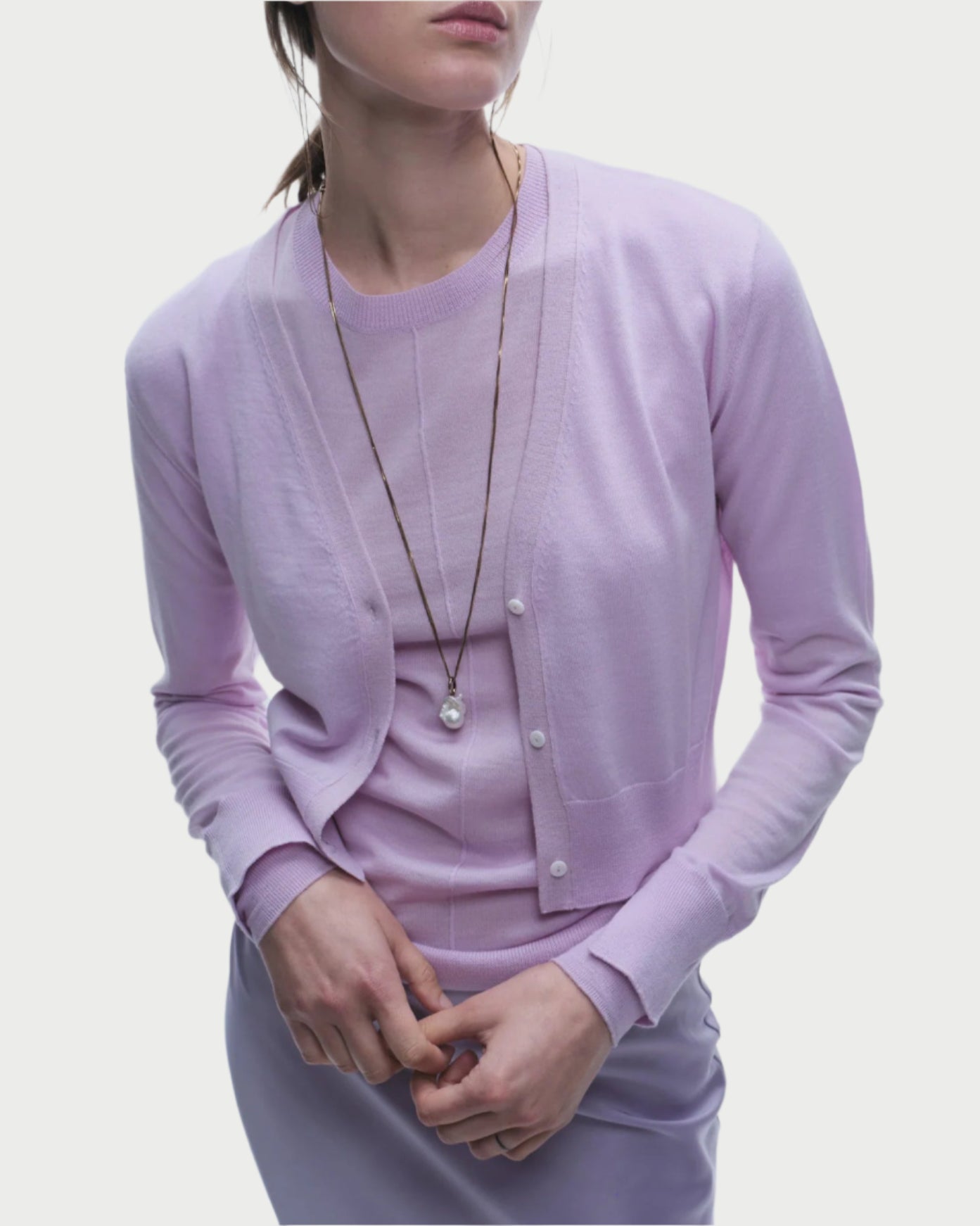 The individual showcases a Saint Art NY Nixon Cropped Cardigan in light purple luxury knitwear, with long sleeves and a round neckline over a matching top. A pendant necklace enhances the elegant, coordinated look against the plain white backdrop.