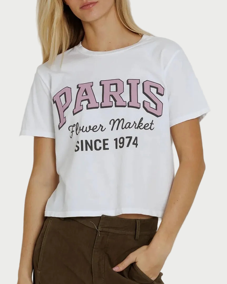 A woman wears the Sunny Somewhere Paris Flower Market Angel Tee—a white, boxy-fit top by Sunny Somewhere featuring “PARIS Flower Market SINCE 1974” printed in pink and black across the front. She pairs the tee with dark pants and her light hair accents the relaxed, effortlessly stylish vibe of her outfit.